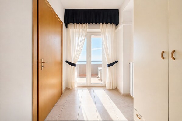 Ein Flur mit Holztür, schwarzem Vorhang, weißen Vorhängen, einer Glasschiebetür, die auf einen Balkon führt, und einem Fliesenboden.