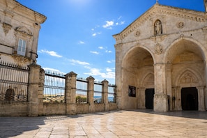 Edifici històric amb una entrada apuntada, mur de pedra amb una tanca metàl·lica i un cel serè.