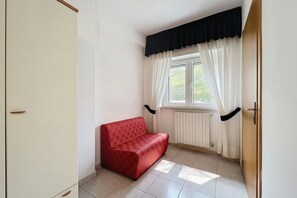 Ein Zimmer mit einem roten Sofa, einem Fenster mit weißen Vorhängen, einem Heizkörper und einer Tür.