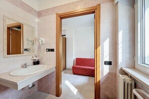 Ein Badezimmer mit Waschbecken, Spiegel und Haartrockner, sowie ein rotes, gemustertes Sofa im angrenzenden Raum.