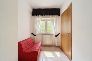 Ein Zimmer mit einer roten Couch, einem Fenster mit weißen Vorhängen, einem Heizkörper und einer Holztür.
