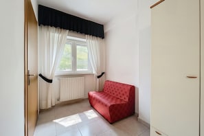Ein Zimmer mit einem roten Sofa, einem Fenster mit weißen Vorhängen, einem Heizkörper und einem Kleiderschrank.