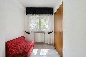 Une pièce avec un canapé rouge, une fenêtre avec des rideaux blancs, un radiateur et une porte en bois.