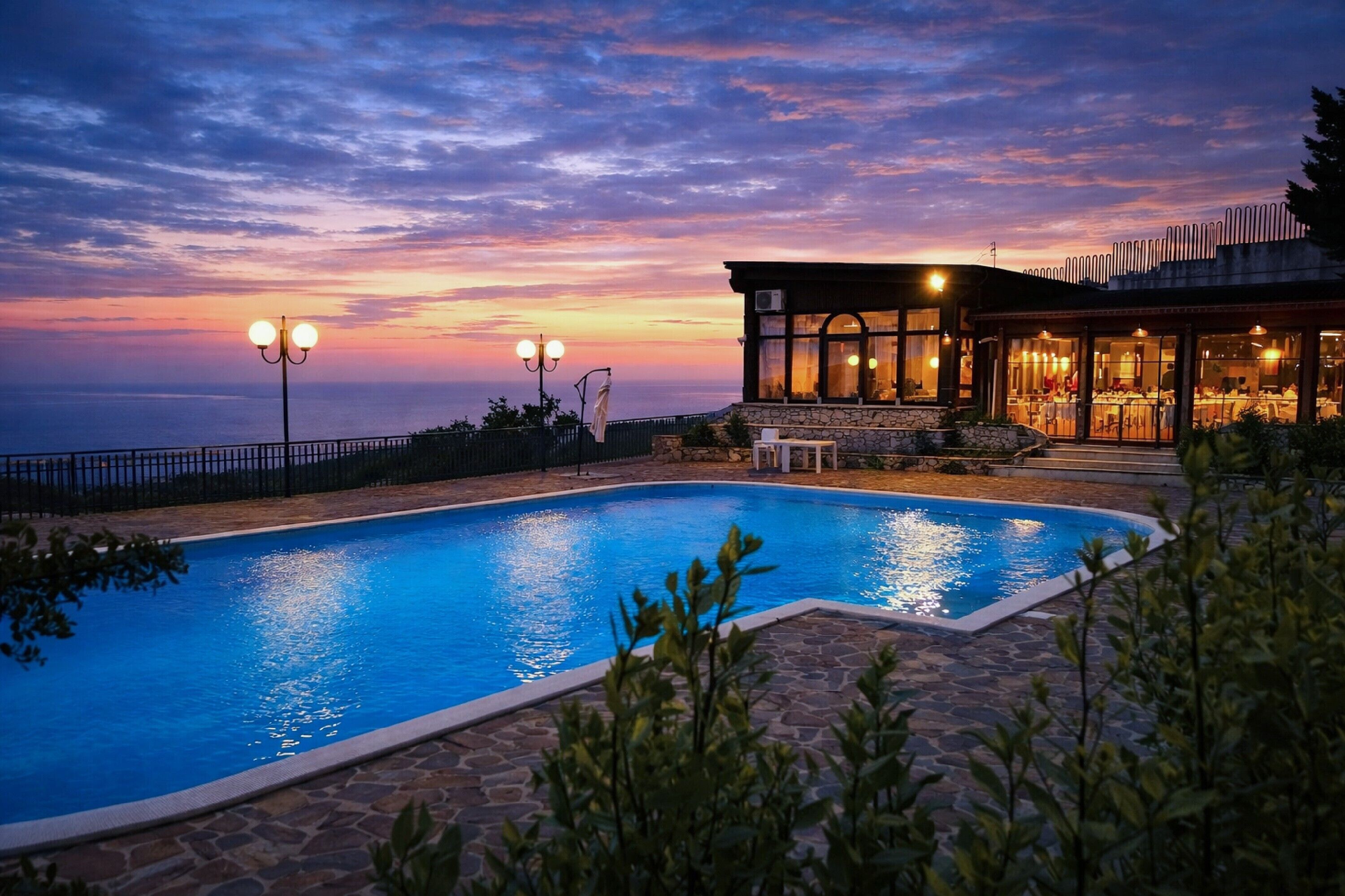 Une piscine aux carreaux de pierre, un bâtiment doté de grandes fenêtres et un ciel au coucher du soleil.