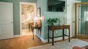 Un couloir avec un plancher en bois, une porte blanche, un téléviseur fixé au mur, une petite table avec une lampe et une plante décorative.