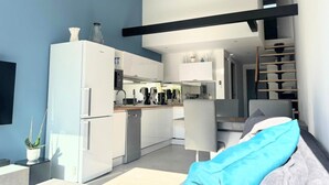 A modern kitchen with white appliances, a dining area with a sofa, and a staircase leading to an upper level. - Naturiste Cap d'Agde Villa Héliovillage 90 (Agde)