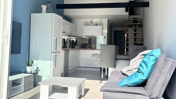 A modern living space with a sofa, coffee table, and a kitchen area visible through a glass door. - Naturiste Cap d'Agde Villa Héliovillage 90 (Agde)