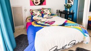 Ein Schlafzimmer mit einem Bett, einer blauen Wand, einem Gemälde und einem kleinen Tisch mit einer Vase.