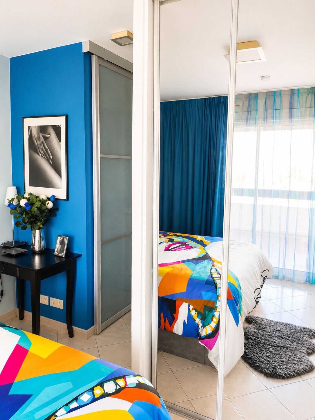 Ein modernes Hotelzimmer mit blauer Akzentwand, einem Bett mit farbenfroher Steudecke, einem Schreibtisch mit Blumenvase und einem großen Fenster mit Vorhängen.