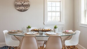 A dining area with a round glass table, white chairs, a wooden clock, and a window with a view.