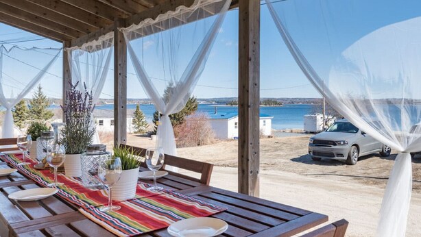 An outdoor dining area with a wooden table set for a meal, overlooking a body of water and a parked car.