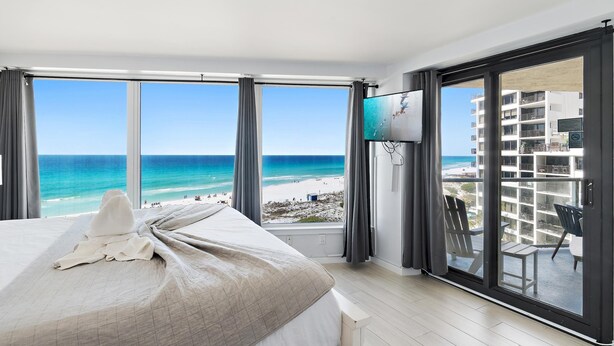 Ein Hotelzimmer mit einem großen Bett, einem Fernseher und einem Balkon mit Blick auf Strand und Meer.