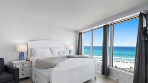 Ein Schlafzimmer mit Bett, Nachttischen, einem Sessel und einem großen Fenster mit Blick auf Strand und Meer.