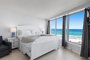 Une chambre à coucher comprenant un lit, des tables de chevet, une chaise et une grande fenêtre donnant sur la plage et l’océan.