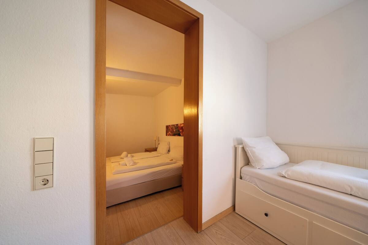 A bedroom with a bed, a bedside table, a wall socket, and a door leading to a bathroom.