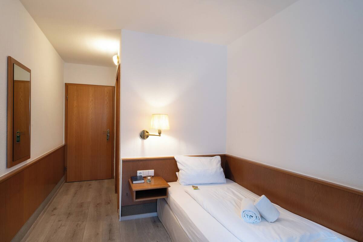 A hotel room with a wooden door, a bedside table with a lamp, a bed with white linens, and a small shelf.