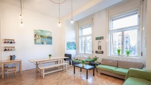 A spacious living room with wooden flooring, a green sofa, a wooden table, and a bench. There are large windows with city views and a painting on the wall. - Spicy Hostel (Budapest)