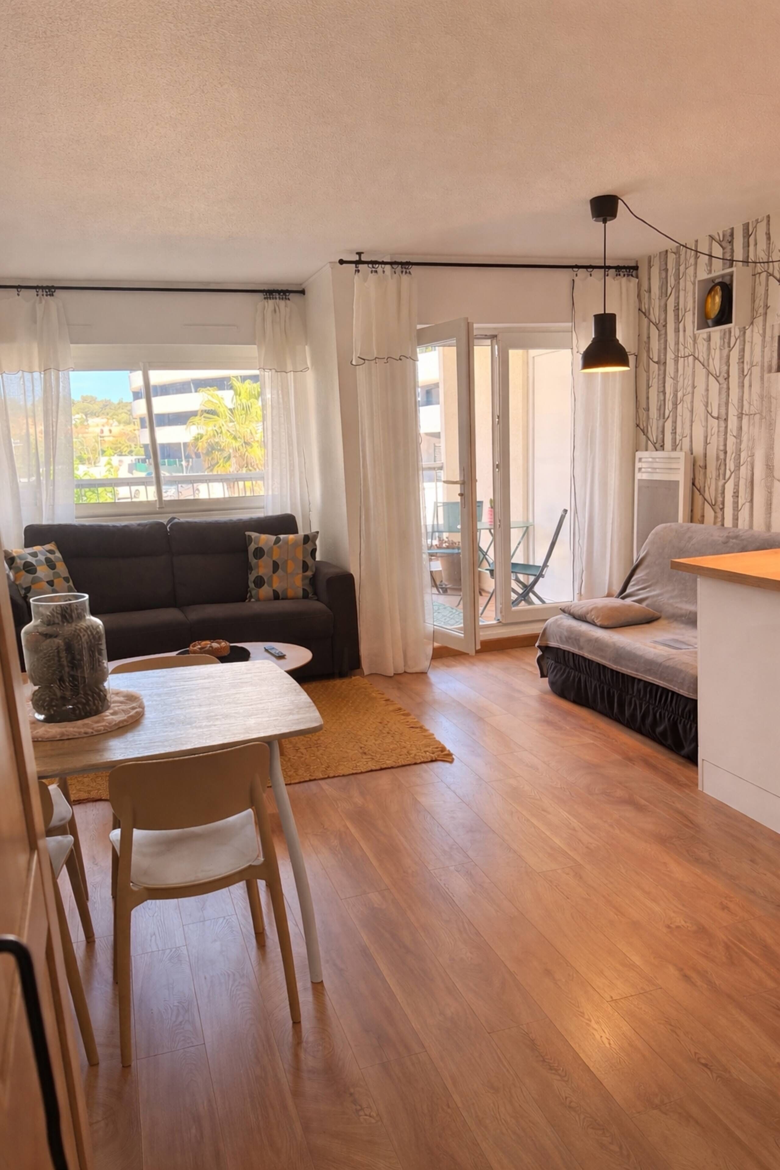 Una sala de estar moderna con un sofá, una mesa de comedor con sillas, una pequeña cocina y un ventanal corredero que da a un espacio exterior.