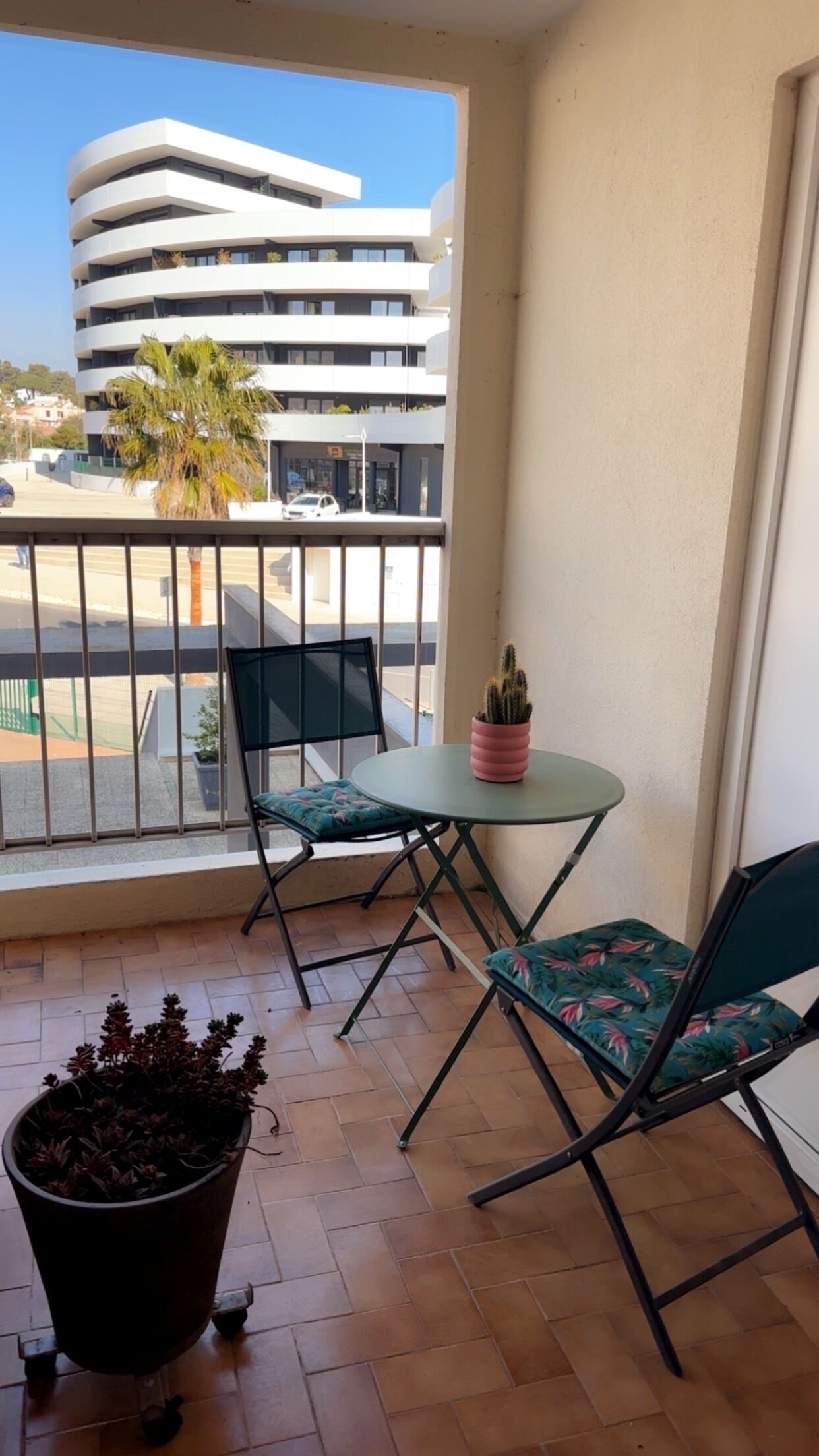 Un balcón con una mesa, sillas, una planta en maceta y vista a un edificio moderno.
