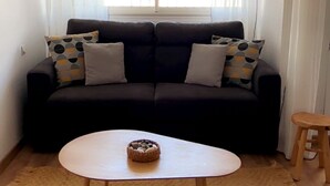 Un salon avec un canapé foncé, une petite table ronde, un tapis beige et une fenêtre ornée de rideaux légers.