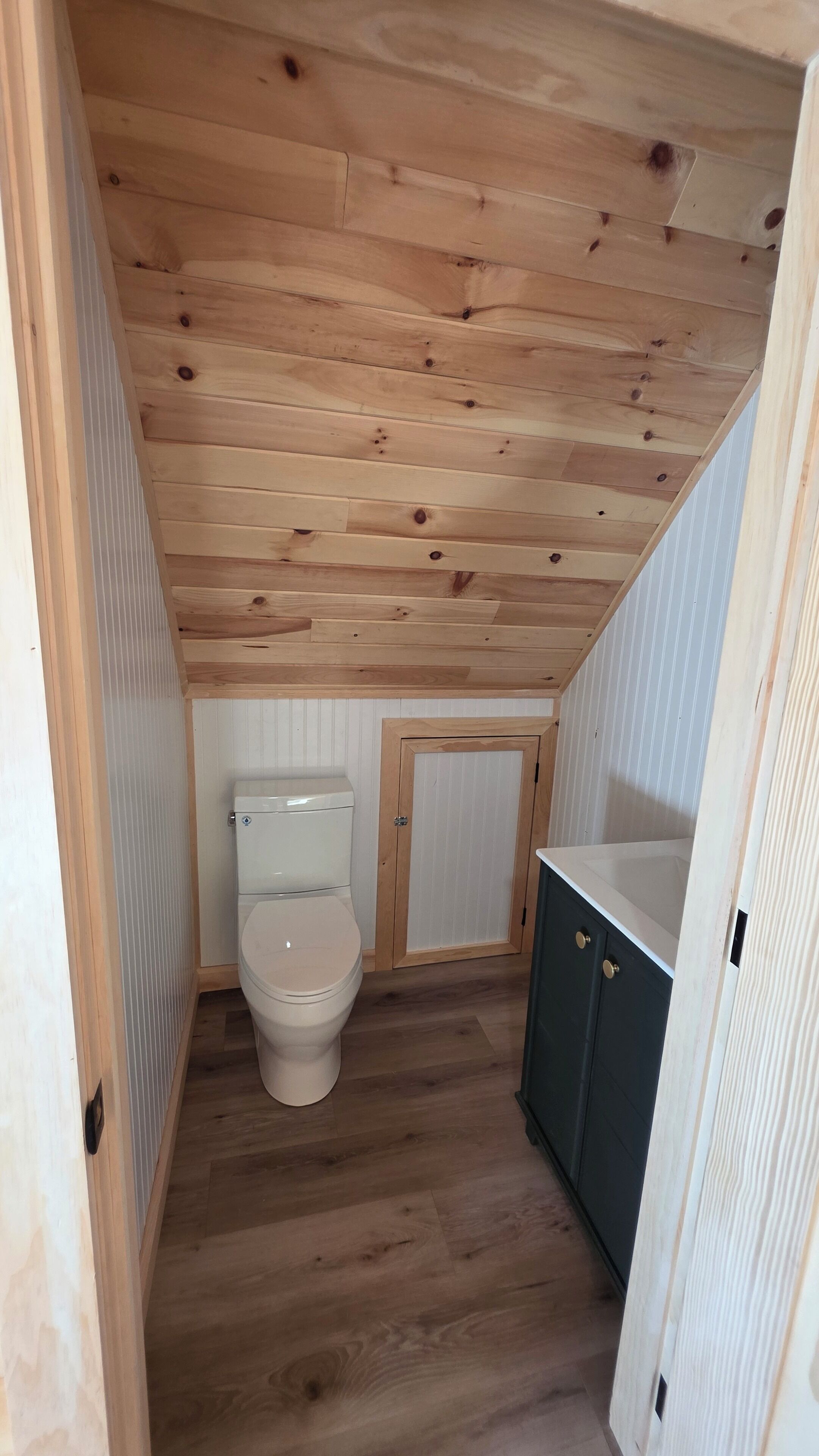 A small, narrow bathroom with a wooden ceiling, a toilet, and a sink with cabinets.