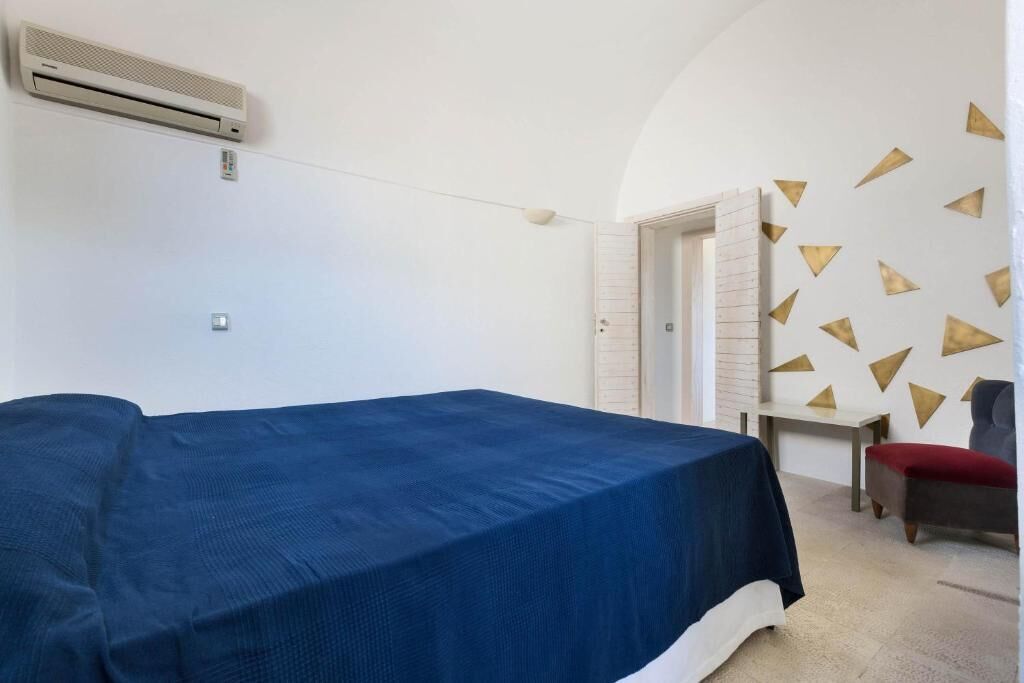 A room with a bed, a chair, and a desk. There is an air conditioning unit on the wall.