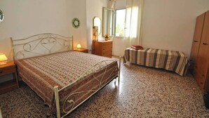 Ein Schlafzimmer mit Metallbettgestell, einem Holzkleiderschrank, einem kleinen Nachttisch mit Lampe, einem Fenster mit Vorhängen und einem gemusterten Teppich.