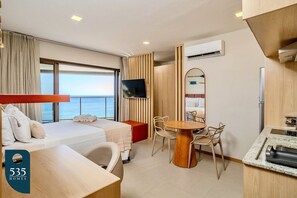 A hotel room with a bed, a TV, a dining area with chairs, a small table, and a view of the sea. - Exclusive ocean view in the heart of Barra (Salvador)