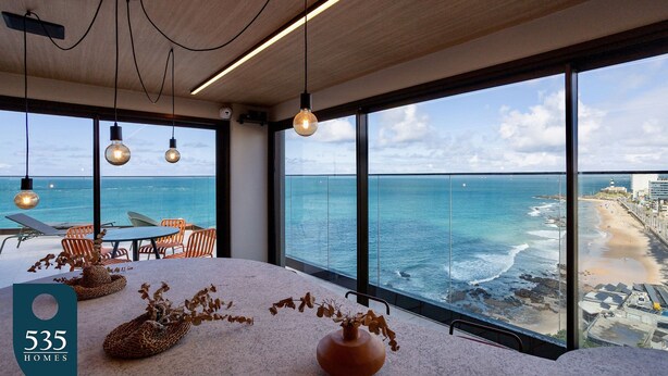Et moderne interiør med store vinduer ud til en strand, et bord med stole og en vase med tørrede planter.