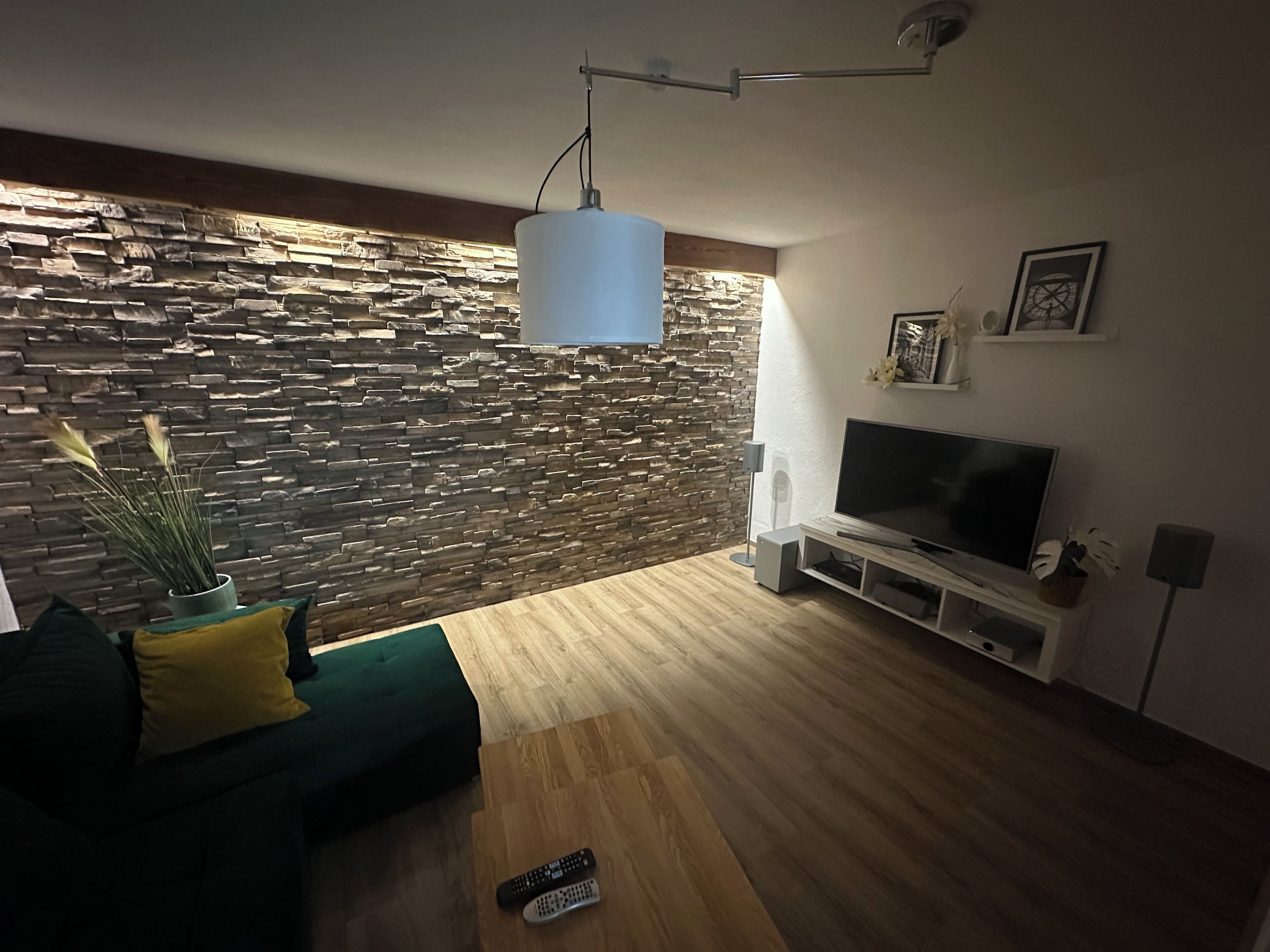 Un saló modern amb una paret de pedra, un sofà verd, una taula de centre de fusta, una televisió i un llum penjant.
