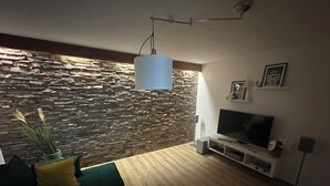 A modern living room with a stone wall, a green sofa, a wooden coffee table, a TV, and a mounted light fixture. - Vacation home Haus zur Böhme (gateway to the Thuringian Forest) (Georgenthal)