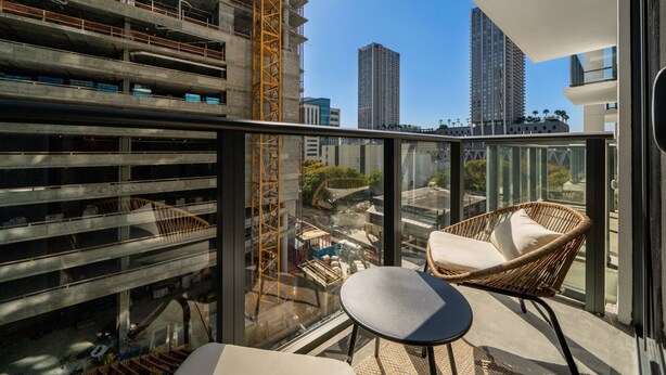 Een balkon met een rieten stoel en een klein tafeltje, met uitzicht op een bouwterrein en hoogbouw.