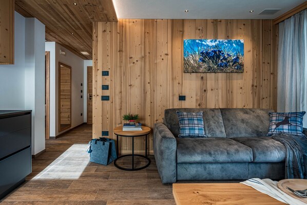 Un salón moderno con un sofá gris, paneles de madera, una mesita redonda con una planta y un cuadro.