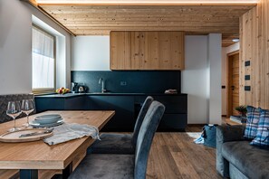 A modern kitchen with wooden cabinets, a dining area with a table set for two, and a sofa in a living area. - 'Genzianella Apartment' with Mountain View, Wi-Fi and Air Conditioning (Cavedago)