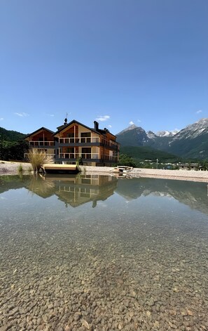 A multi-story building with a clear reflection in a shallow water body, surrounded by mountains and greenery. - 'Pino Mugo' Apartment with Mountain View, Wi-Fi and Air Conditioning (Cavedago)