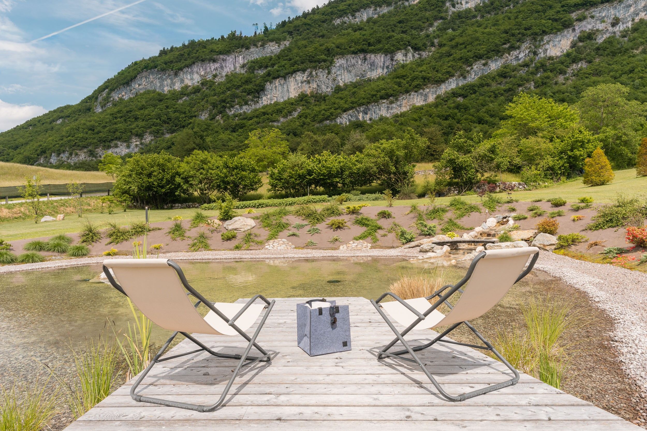 A wooden deck with two lounge chairs facing a calm body of water, surrounded by a natural landscape with a mountain in the background.