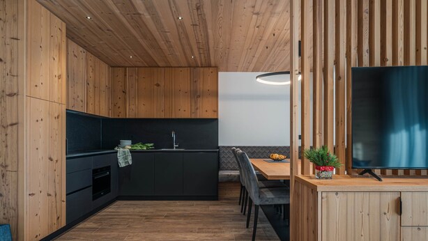 Cozinha moderna com armários de madeira, área de jantar com mesa e cadeiras, e uma TV de tela plana fixada em um suporte de madeira.