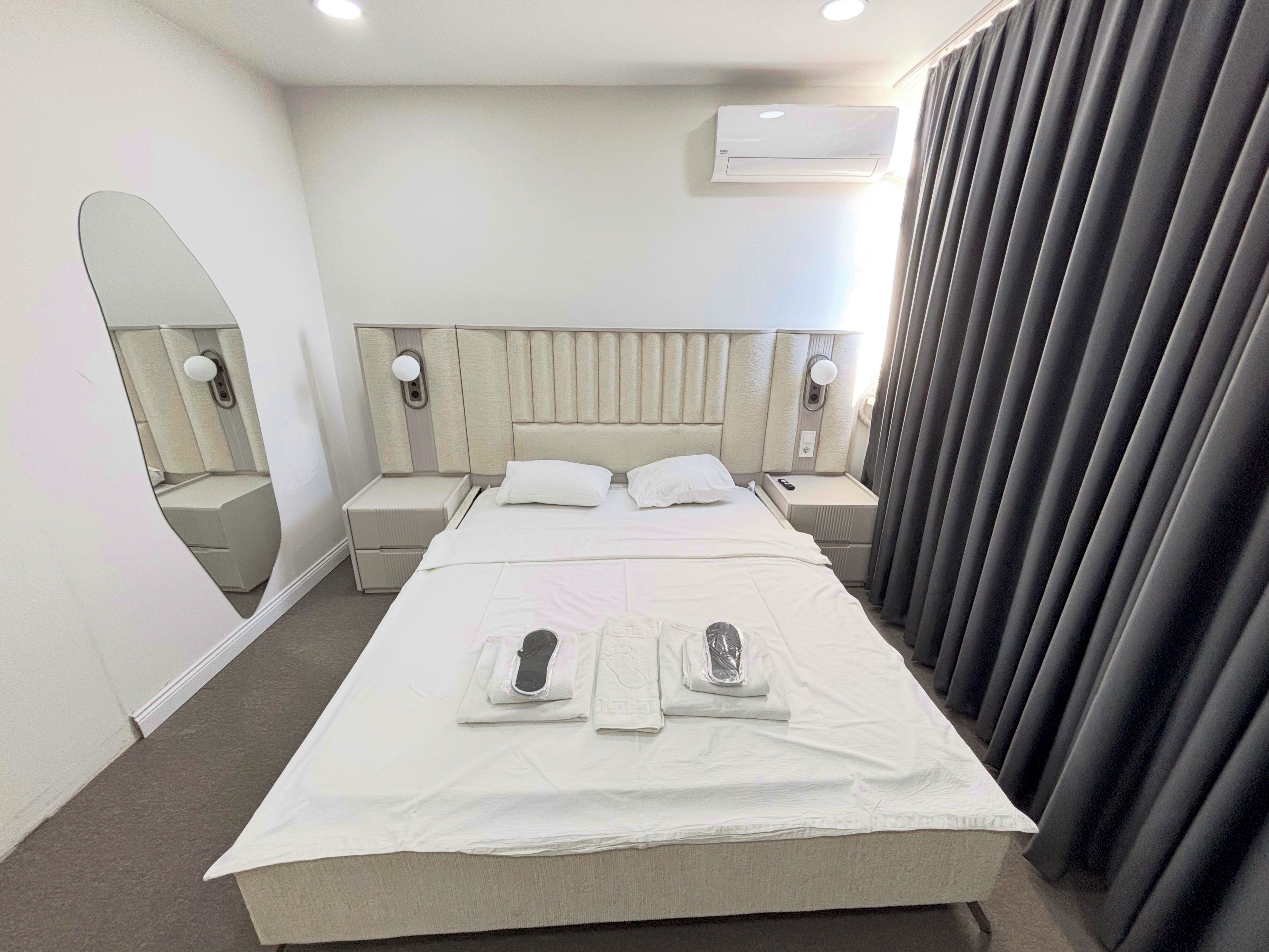A hotel room with a large bed, bedside tables, a mirror, an air conditioning unit, and a window with curtains.