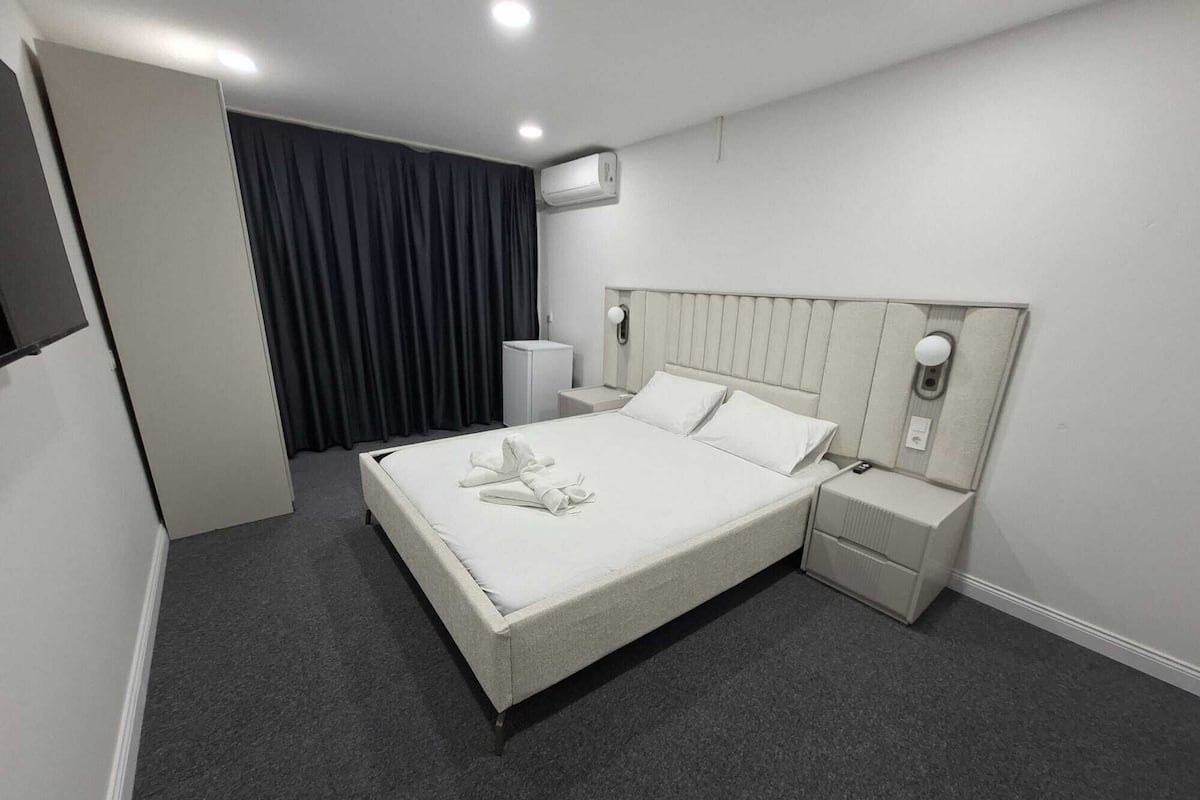 Ett hotellrum med en stor säng, ett sängbord, en liten avlastningsyta, en spegel och ett fönster med gardiner.