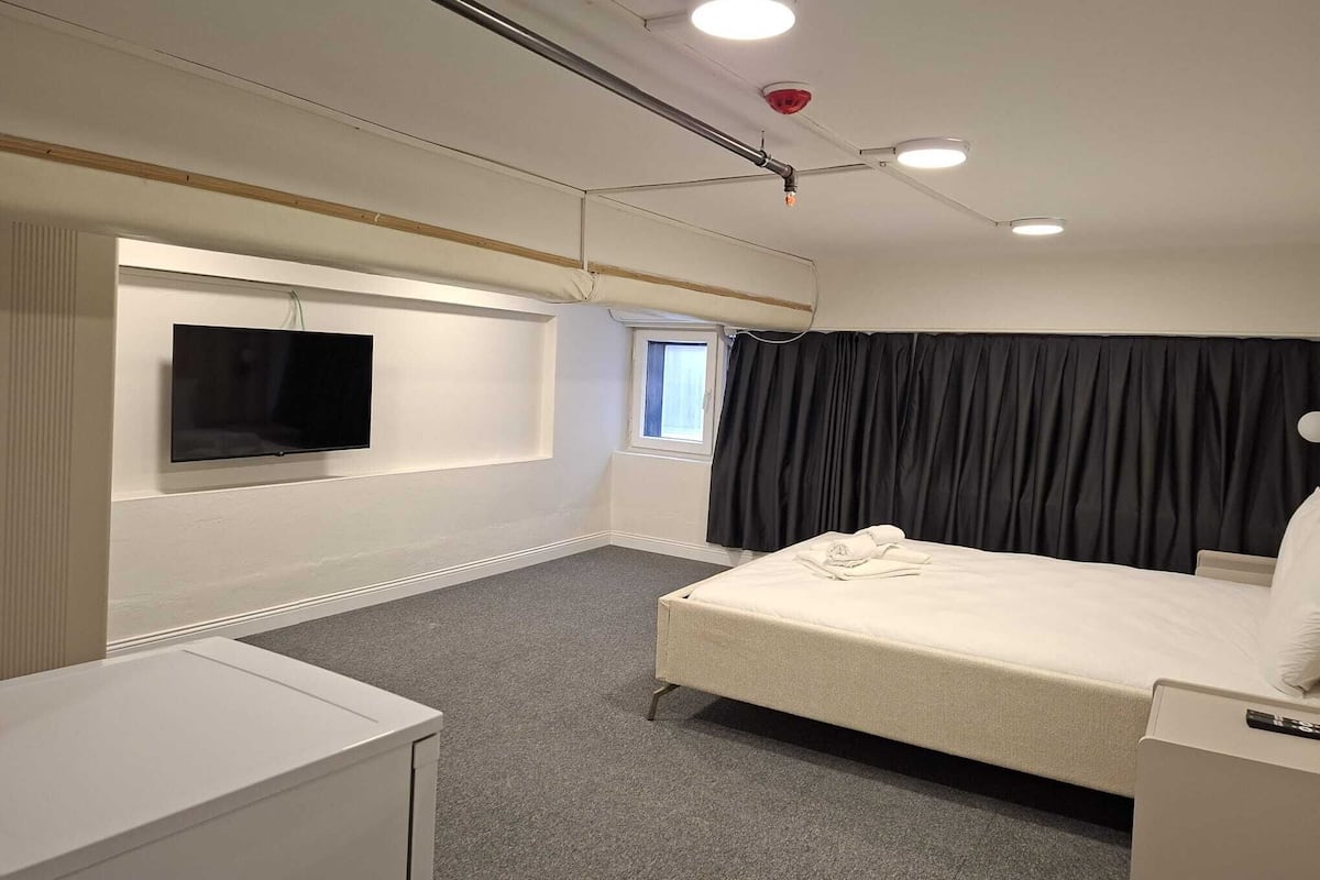 Ett hotellrum med en säng, en tv som är monterad på väggen, ett fönster med gardiner och ett tak med lampor.