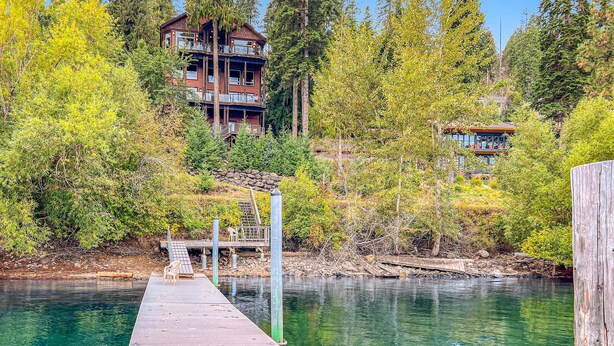 A wooden dock extending into a clear body of water, with a multi-story building in the background surrounded by trees. - Unwind at Deja View Upper (Hayden Lake)