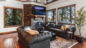 A modern living room with a stone fireplace, leather sofas, and large windows offering views of trees. - Unwind at Deja View Upper (Hayden Lake)