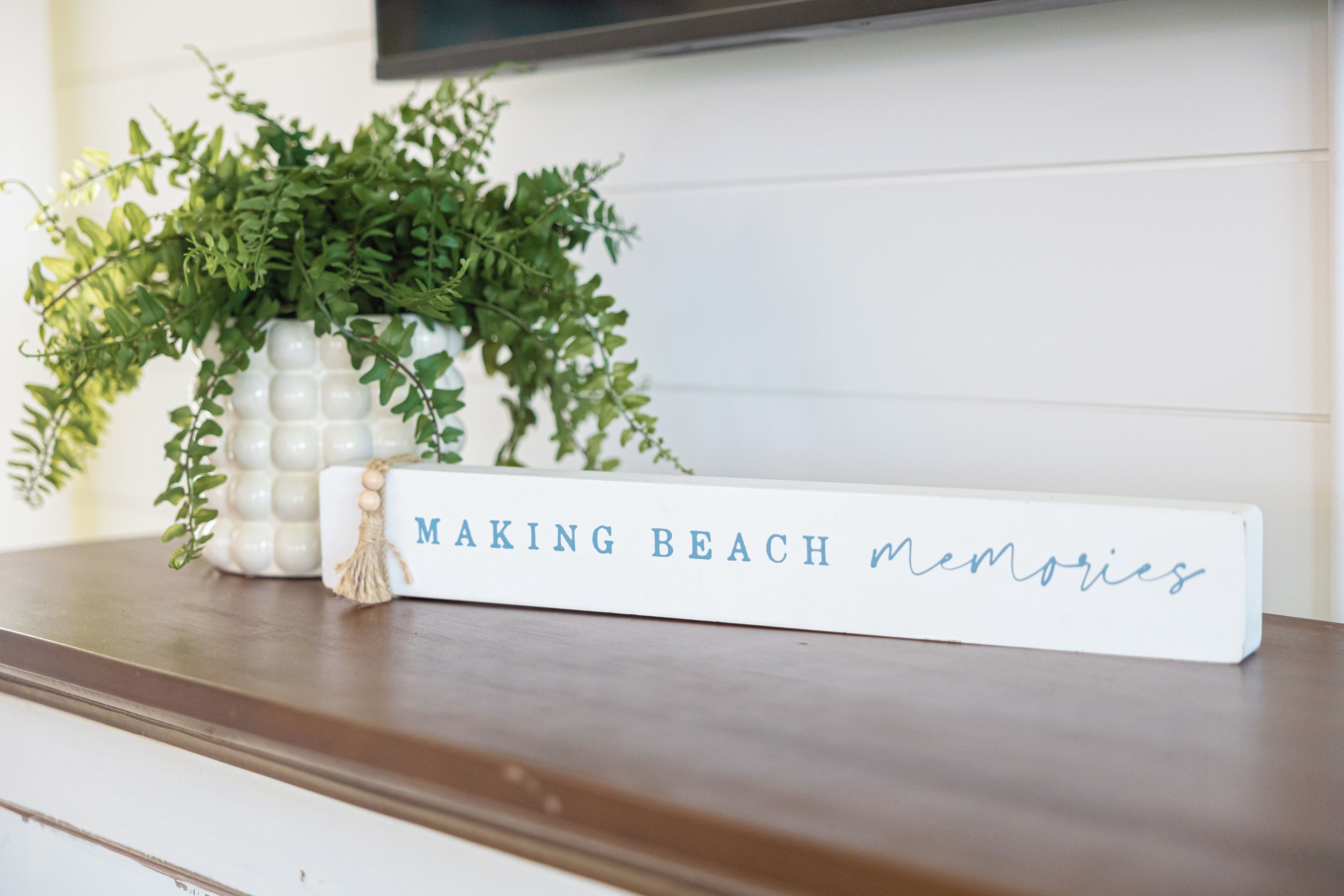 Un vase décoratif blanc, orné de feuillage vert, est posé sur une surface en bois, à côté d’un panneau sur lequel on peut lire : « Créez des souvenirs inoubliables à la plage ».