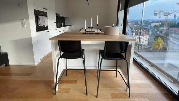 A modern kitchen with a wooden island, two black chairs, and a large window offering a city view. - Apt near City Center, Novartis & Messe+Basel Card (Basel)