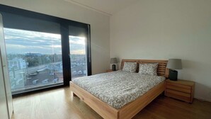 A bedroom with a large window, a wooden bed, two bedside tables with lamps, and a view of a cityscape. - Apt near City Center, Novartis & Messe+Basel Card (Basel)