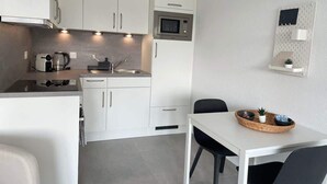 Kompakti keittiö, jossa on valkoiset kaapit, mikroaaltouuni, vedenkeitin ja pieni ruokailutila, jossa on pöytä ja tuolit.