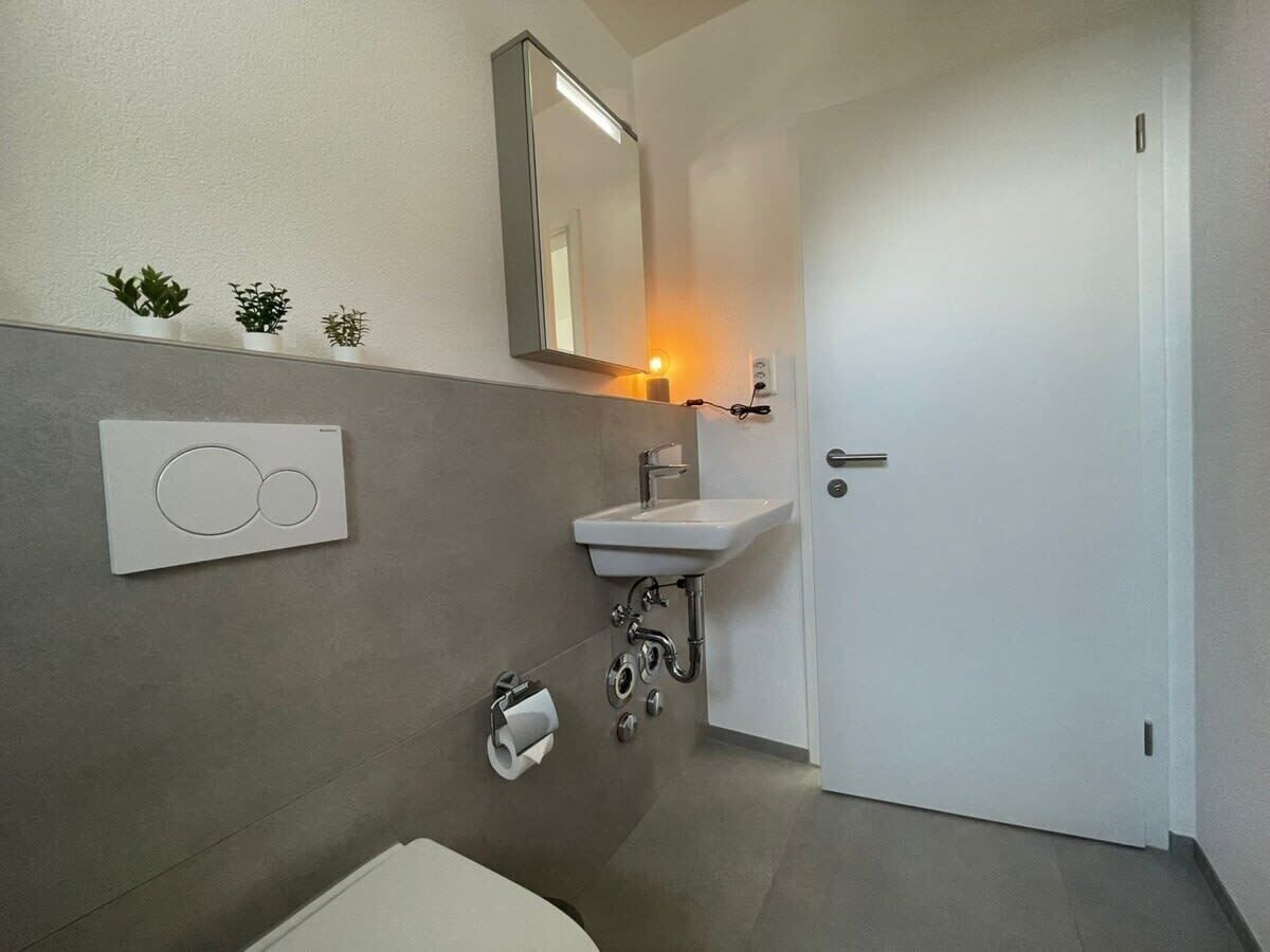 A modern bathroom with a white sink, mirror, and toilet. There are three small potted plants on the wall and a light fixture above the mirror.