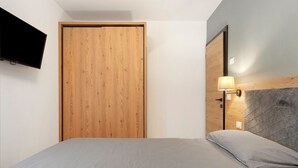 A bedroom with a wooden wardrobe, a bed with grey bedding, a mounted TV, and a wall-mounted lamp.