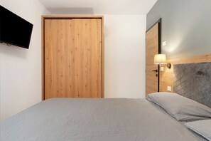 Quarto com guarda-roupa de madeira, cama com roupa de cama cinza, TV fixada na parede e luminária também fixada na parede.