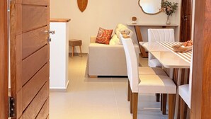 A modern interior with a wooden door, a dining area with a white table and benches, a sofa with cushions, a hanging light fixture, and a small stool. - Apartamento Messina  (Jijoca de Jericoacoara)